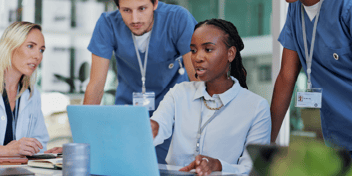 A group of healthcare professionals, including a woman in business attire and three medical staff in blue scrubs, gathered around a laptop in a modern office setting. The woman in business attire is leading the discussion, pointing at the screen, while the others attentively engage in the conversation.