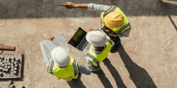 Three construction professionals wearing yellow and white hard hats and high-visibility vests stand on a construction site. They are reviewing blueprints and using a laptop while one of them points in a direction, likely discussing project details. The overhead shot captures the collaborative nature of their work.
