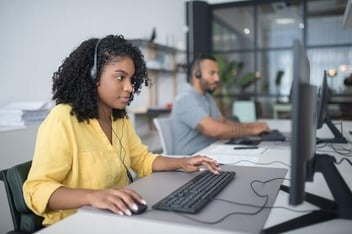 A woman in a yellow shirt sits at a desk wearing a headset and using a computer mouse, while a male colleague works in the background of a bright, open-plan office.